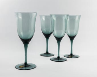 Puede incluir: Un juego de cuatro copas de vino con tallo de cristal azul pizarra translúcido. Cada copa presenta un cuenco ancho y acampanado que se estrecha hacia un tallo delgado y una base circular. Una pequeña pegatina dorada es visible en la base.