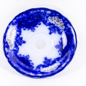 May include: A vintage blue and white porcelain saucer with a scalloped edge. The saucer features a detailed floral and scroll design in blue against a white background. Gold accents are present in the center and around the rim.