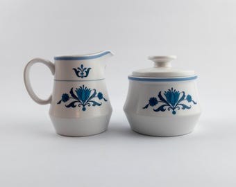 Puede incluir: Una lechera y un azucarero con tapa de cerámica blanca, ambos con un patrón floral azul y una fina banda azul cerca del borde superior. La lechera tiene un asa curva y un pico vertedor, mientras que el azucarero incluye una tapa a juego.