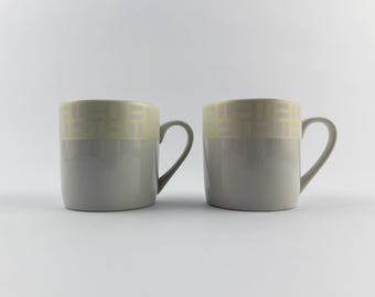 Puede incluir: Dos tazas de cerámica con un acabado liso gris claro en la parte inferior y una banda ancha alrededor del borde superior con un patrón geométrico repetitivo de rectángulos de color amarillo claro y gris. Cada taza tiene un asa curva.