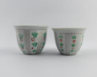 Puede incluir: Dos tazas de cerámica blanca con un patrón repetitivo de palmeras verdes estilizadas y pequeños símbolos rojos del zodiaco Tauro. Las tazas tienen paneles verticales texturizados con un diseño gris claro similar a escamas.