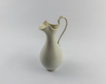 Puede incluir: Una jarra de cerámica de color crema con un acabado liso y brillante. El recipiente presenta una base bulbosa, un cuello esbelto y un pico vertedor curvado y ensanchado. Un asa única, delgada y arqueada, está fijada al lado del cuello.