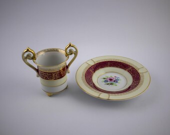 Puede incluir: Un juego de taza de dos asas y platillo de porcelana con base color crema, una banda de color burdeos intenso y detalles de filigrana dorada. El centro del platillo muestra un pequeño motivo floral pintado a mano en rosa y azul.