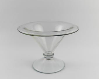 Puede incluir: Un cuenco de cristal transparente sobre pedestal con una base ancha y poco profunda y un borde liso y redondeado. El cuenco se apoya en un tallo fino y cónico que se conecta a una base circular plana, creando un diseño equilibrado.