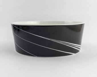 Puede incluir: Un cuenco redondo de cerámica con un exterior negro brillante que presenta finas líneas diagonales blancas. El interior del cuenco es de color blanco sólido y el borde es fino y blanco, creando un contraste marcado con la superficie oscura.