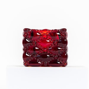 May include: A square, ruby-red glass candle holder with a quilted pattern. The design features rounded shapes and small, raised dots, creating a textured surface. The candle holder is a decorative item.