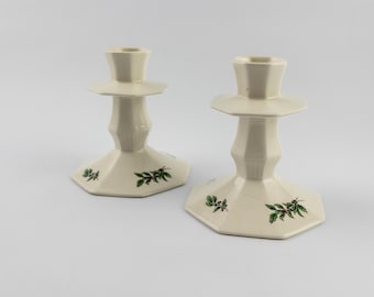 Puede incluir: Dos candelabros de cerámica en color blanco roto con base octogonal y parte superior ensanchada. Cada candelabro presenta un diseño pintado de hojas de acebo verdes y pequeñas bayas rojas alrededor de la base. Los tallos son facetados.