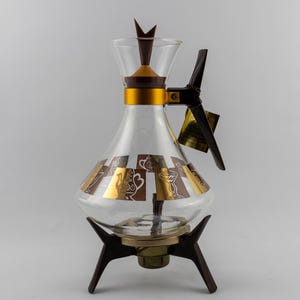 May include: A clear glass carafe with a gold metal neck band and a dark brown handle. The body features a repeating pattern of brown and gold squares with teapot illustrations. It rests on a dark brown tripod stand with a central metal base.