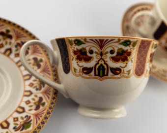 Puede incluir: Un juego de taza de té y platillo de cerámica blanca con un patrón decorativo en tonos óxido, azul marino, verde y dorado. La taza tiene un asa curva y una base con pie, con un diseño floral y geométrico que rodea el exterior.