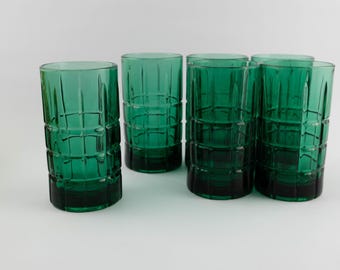 Puede incluir: Un conjunto de cinco vasos cilíndricos hechos de vidrio verde esmeralda translúcido. Cada vaso presenta un patrón de cuadrícula repetitivo de líneas verticales y horizontales en relieve en la superficie exterior, creando un diseño geométrico texturizado.