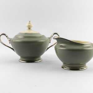 May include: A sage green sugar bowl with a lid and a matching creamer pitcher. Both pieces feature a rounded body, a pedestal base, and metallic silver-toned handles and rims. The sugar bowl lid has a small, cream-colored finial on top.