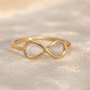 May include: A gold ring with an infinity symbol design. The ring's delicate form is set against a soft, neutral background. The infinity symbol is a continuous loop, symbolising eternity. The ring is likely to be worn on a finger.