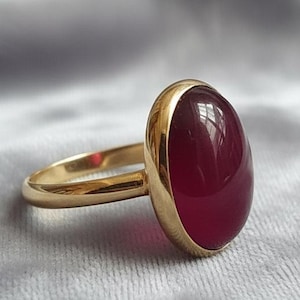 May include: A gold ring featuring a large, oval-shaped, deep red gemstone. The gemstone is set in a gold bezel, and the band is a simple, polished gold. The ring is displayed on a soft, gray fabric.