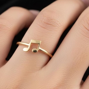 以下が含まれることがあります： 金色のリングで、音符のデザインが特徴です。リングは細く繊細で、音符の形がはっきりと定義されています。指に着用されており、ジュエリーのシンプルなエレガンスを表現しています。背景は暗いです。