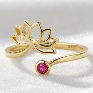 May include: A gold-colored ring featuring a lotus flower design. The ring has an open design with a small, round, red gemstone. The ring is made of a shiny, gold-toned metal.