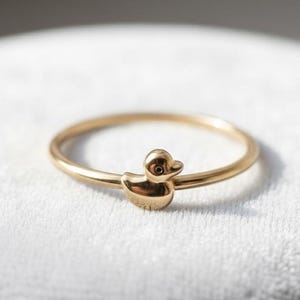 May include: A gold ring with a duck charm. The ring has a slender band and a detailed duck design. The ring is displayed on a white surface. The ring is suitable for everyday wear.