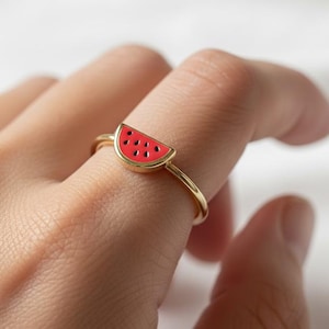 May include: A gold-colored ring featuring a watermelon slice design. The watermelon slice is red with black seeds and a green rind. The ring is worn on a finger, showcasing the jewelry's details.