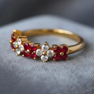May include: A gold ring featuring a floral design. The ring is adorned with alternating red and white gemstones, arranged in a flower pattern around the band. The ring is set on a gray surface.