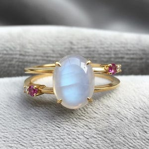 Op de afbeelding: Twee gouden ringen. De bovenste ring heeft een grote, ovale, lichtblauwe maansteen. De onderste ring heeft een kleinere, ovale maansteen en twee kleine roze edelstenen. De ringen zijn geplaatst tegen een zachte, grijze achtergrond.