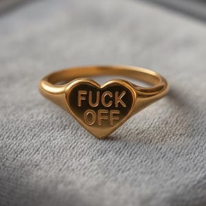 Puede incluir: Anillo dorado con una cara en forma de corazón. El corazón presenta las palabras "FUCK OFF" en mayúsculas. El anillo está sobre un fondo gris texturizado.