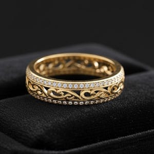 Pode incluir: Um anel dourado com um intrincado design de filigrana. O anel apresenta uma faixa de pequenas pedras preciosas transparentes colocadas entre os padrões de filigrana. O anel é exibido em uma superfície preta.