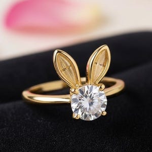 Puede incluir: Un anillo dorado con un diseño de orejas de conejo. El anillo tiene una gema grande, redonda y transparente en el centro. Las orejas de conejo son detalladas y se extienden hacia arriba. El anillo está sobre un fondo negro.