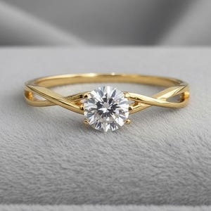 May include: A gold ring with a large, round, clear gemstone. The band features an interwoven design. The ring is set against a soft, gray background, highlighting the sparkle of the gemstone. A classic piece of jewelry.