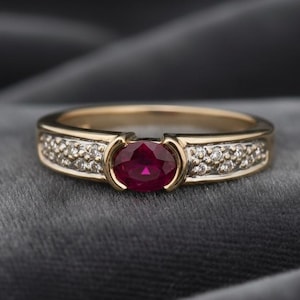 Puede incluir: Un anillo de oro con una gran piedra central de rubí ovalada. La banda está adornada con pequeños diamantes brillantes. El anillo se muestra sobre un fondo gris oscuro, resaltando su brillo y diseño.