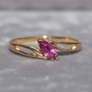 May include: A gold ring featuring a marquise-cut, pink gemstone. The ring's band has an open design on either side of the gemstone. The ring is set against a soft, grey background.