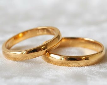 May include: Two gold wedding bands are slightly overlapping on a white, textured surface. The rings are simple, with a smooth, polished finish. The bands are a warm, yellow gold color, reflecting light.