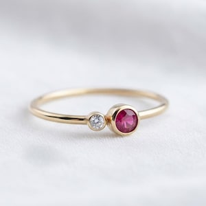 Pode incluir: Um delicado anel de ouro com uma gema vermelha redonda e vibrante e uma gema menor e transparente. A banda fina e o design minimalista do anel criam uma estética elegante e intemporal. As pedras preciosas são definidas em uma configuração de moldura.