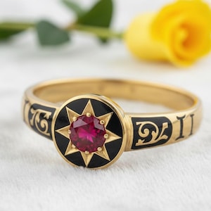 May include: A gold ring with a central red gemstone, surrounded by a black starburst design. The ring band features black enamel detailing. A yellow rose and green leaves are in the background.