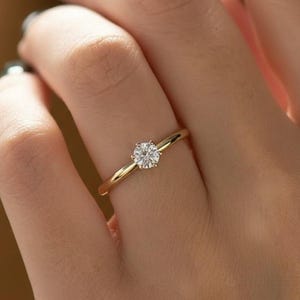 May include: A gold ring with a single, round, clear gemstone. The ring is on a finger, and the band is thin and polished. The gemstone is set in a classic, four-prong setting. The ring is a simple and elegant design.