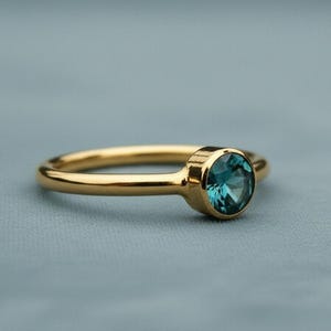 Puede incluir: Un anillo de oro con una gema redonda de color turquesa. El anillo tiene un diseño simple y elegante con una banda lisa y pulida. La gema está engastada en un bisel, lo que realza su color y brillo.