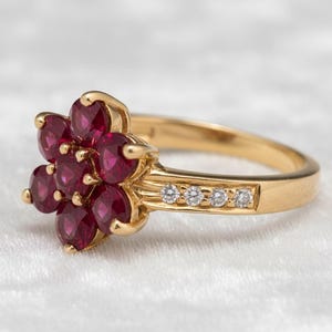 May include: A gold ring featuring a cluster of oval-cut, deep red gemstones arranged in a flower shape. The band is gold, with small, clear gemstones set along the side. The ring is set against a soft, white background.