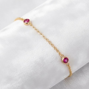 Può includere: Un delicato braccialetto d'oro con tre gemme rosa rotonde e vibranti. La catena del braccialetto è un design sottile e collegato, e le gemme sono incastonate in semplici castoni. Il gioiello poggia su un tessuto bianco e morbido.