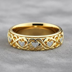 May include: A gold-colored ring with a repeating heart design. The ring features a series of openwork hearts, each with a small cluster of clear stones. The band has a polished finish and sits on a gray surface.
