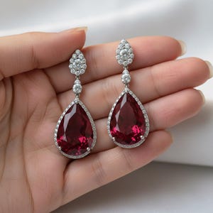 May include: A pair of elegant drop earrings featuring large, pear-shaped, deep red gemstones. The gemstones are surrounded by a halo of small, sparkling clear stones. The earrings have a delicate, silver-toned setting and are designed for pierced ears.