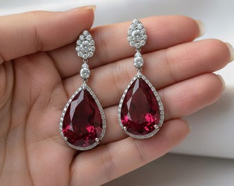 May include: A pair of drop earrings with large, pear-shaped, deep red gemstones. The gemstones are framed by a halo of small, sparkling clear stones. The earrings have a delicate, silver setting and are designed for pierced ears.