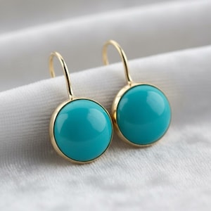 May include: A pair of gold-coloured earrings featuring a round, bright turquoise stone. The earrings have a simple, elegant design, with a gold frame around the turquoise stone. The earrings are displayed on a white, textured surface.