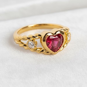 May include: A gold ring with a heart-shaped, red gemstone center. The ring features a twisted rope design on the band and is accented with two smaller, round, clear gemstones on either side of the heart-shaped stone. The ring is set against a white background.