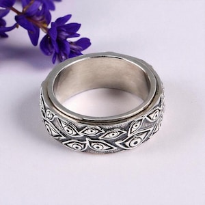 May include: A silver-colored spinner ring with an intricate design of stylized eyes. The ring has a rotating central band and is set against a soft, neutral background with a sprig of purple flowers.