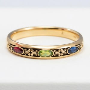 Puede incluir: Un anillo de banda dorado con tres piedras preciosas ovaladas: roja, verde y azul. El anillo presenta un dise&ntilde;o ornamentado con volutas entre las piedras preciosas. El anillo est&aacute; sobre un fondo blanco.