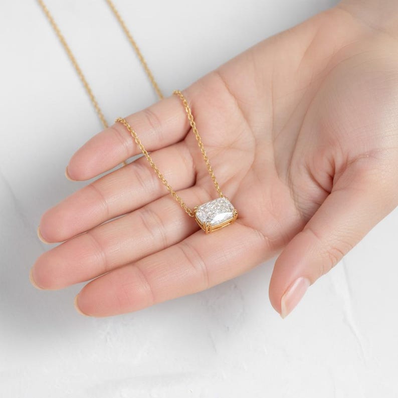 K&ouml;nnte beinhalten: Eine goldfarbene Halskette mit einem rechteckigen Anh&auml;nger, besetzt mit klaren Steinen. Die Kette wird in einer Hand gehalten, um das Design des Anh&auml;ngers hervorzuheben. Die Kette ist fein, und der Anh&auml;nger ist etwa 1,5 cm lang. Das Gold unterstreicht das Funkeln der Steine.