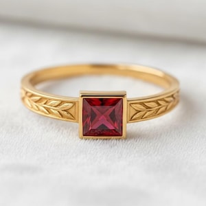 May include: A gold ring with a square, red gemstone. The band features a leaf design. The gemstone is set in a square bezel. The ring is on a white surface.