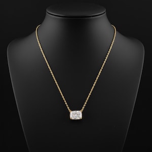 K&ouml;nnte beinhalten: Eine goldene Halskette mit einem rechteckigen, klaren Edelsteinanh&auml;nger. Der Anh&auml;nger ist in einer goldenen L&uuml;nette gefasst und h&auml;ngt an einer feinen Goldkette. Die Halskette wird auf einer schwarzen Schaufensterpuppe pr&auml;sentiert.