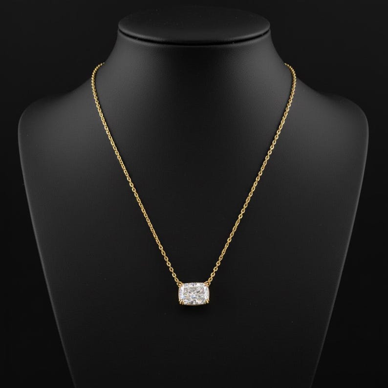 K&ouml;nnte beinhalten: Eine goldene Halskette mit einem rechteckigen, klaren Edelsteinanh&auml;nger. Der Anh&auml;nger ist in einer goldenen L&uuml;nette gefasst und h&auml;ngt an einer feinen Goldkette. Die Halskette wird auf einer schwarzen Schaufensterpuppe pr&auml;sentiert.