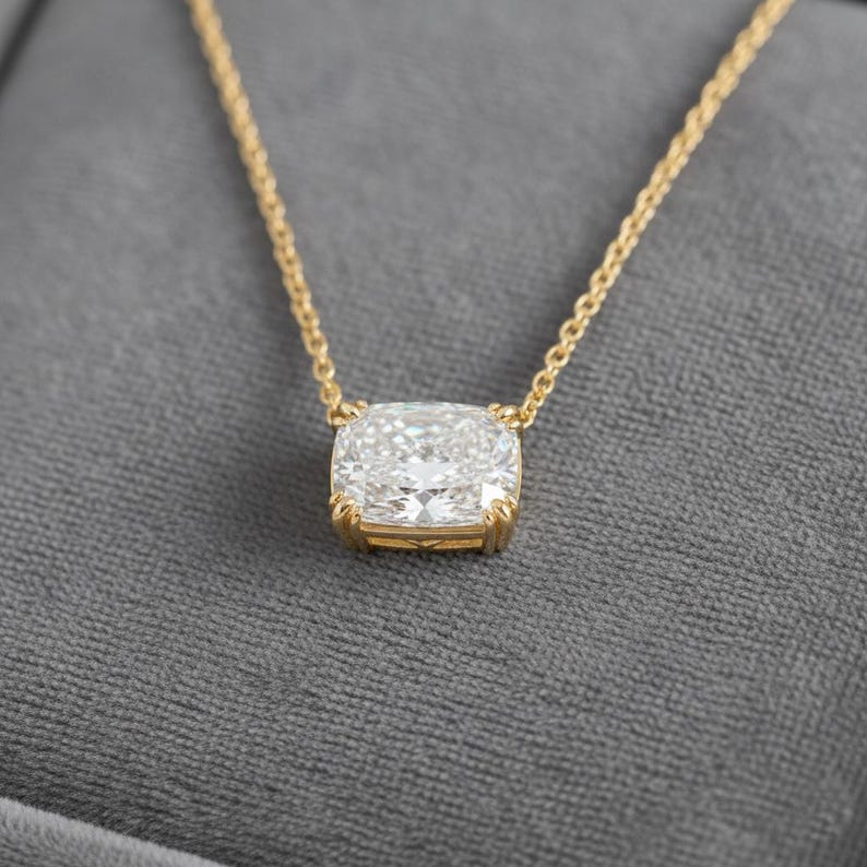 K&ouml;nnte beinhalten: Eine goldene Halskette mit einem gro&szlig;en, quadratisch geschliffenen Diamantanh&auml;nger. Der Diamant ist in einer goldenen L&uuml;nette gefasst und h&auml;ngt an einer zarten Goldkette. Der Hintergrund ist ein weicher grauer Stoff.