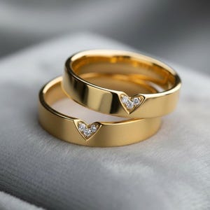 May include: Two gold rings, one on top of the other. Each ring features a heart-shaped recess set with small, clear gemstones. The rings have a smooth, polished finish and are displayed against a soft, grey background.