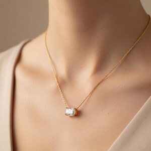 K&ouml;nnte beinhalten: Eine goldene Halskette mit einem rechteckigen, klaren Edelsteinanh&auml;nger. Der Anh&auml;nger ist in einer goldenen L&uuml;nette gefasst und h&auml;ngt an einer zarten Goldkette. Die Halskette wird am Hals einer Person getragen.